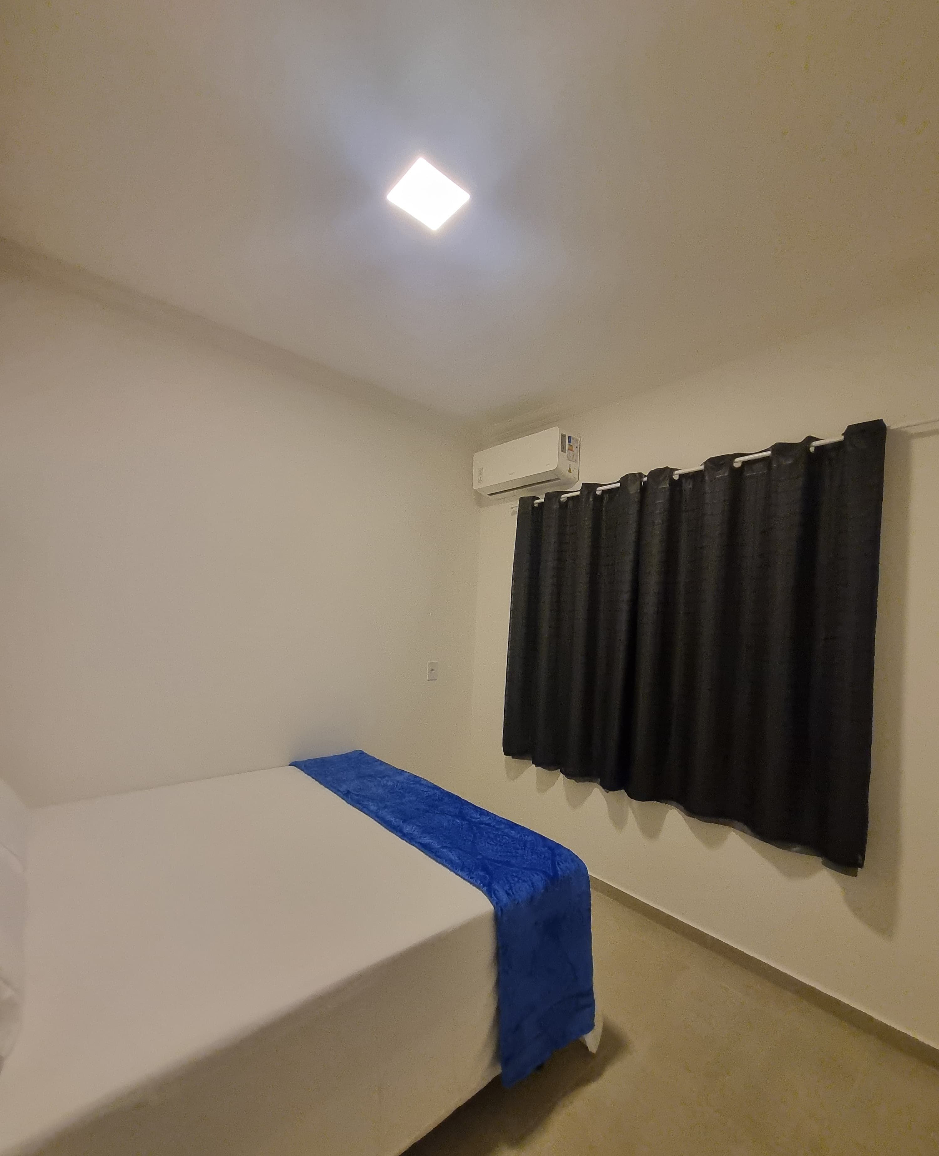 A single bed with a blue bedspread in a room with a white ceiling and a black curtain.