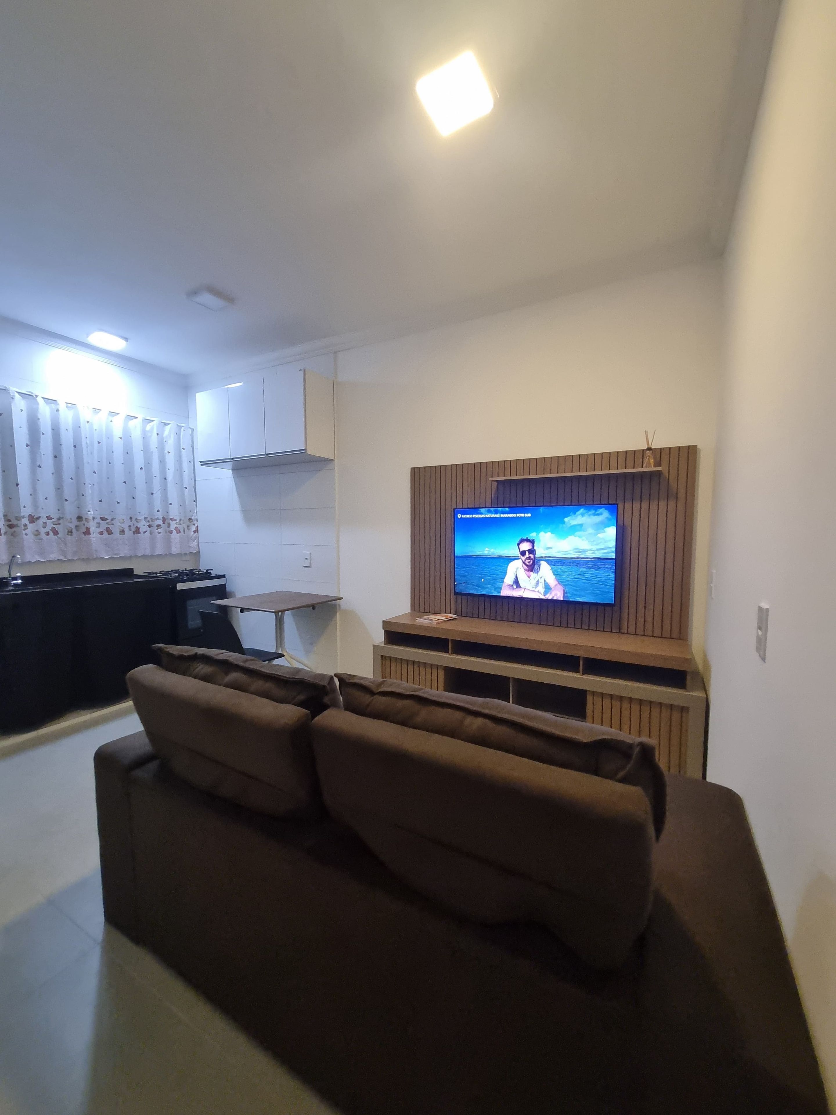 Una sala de estar con un televisor de pantalla plana sobre un mueble de madera, un sofá marrón y una cocina al fondo.