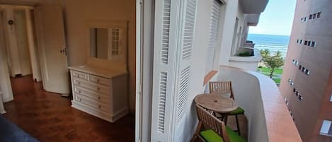 A balcony with a view of the sea, a wooden chair with a green cushion, a white dresser, and a mirror.