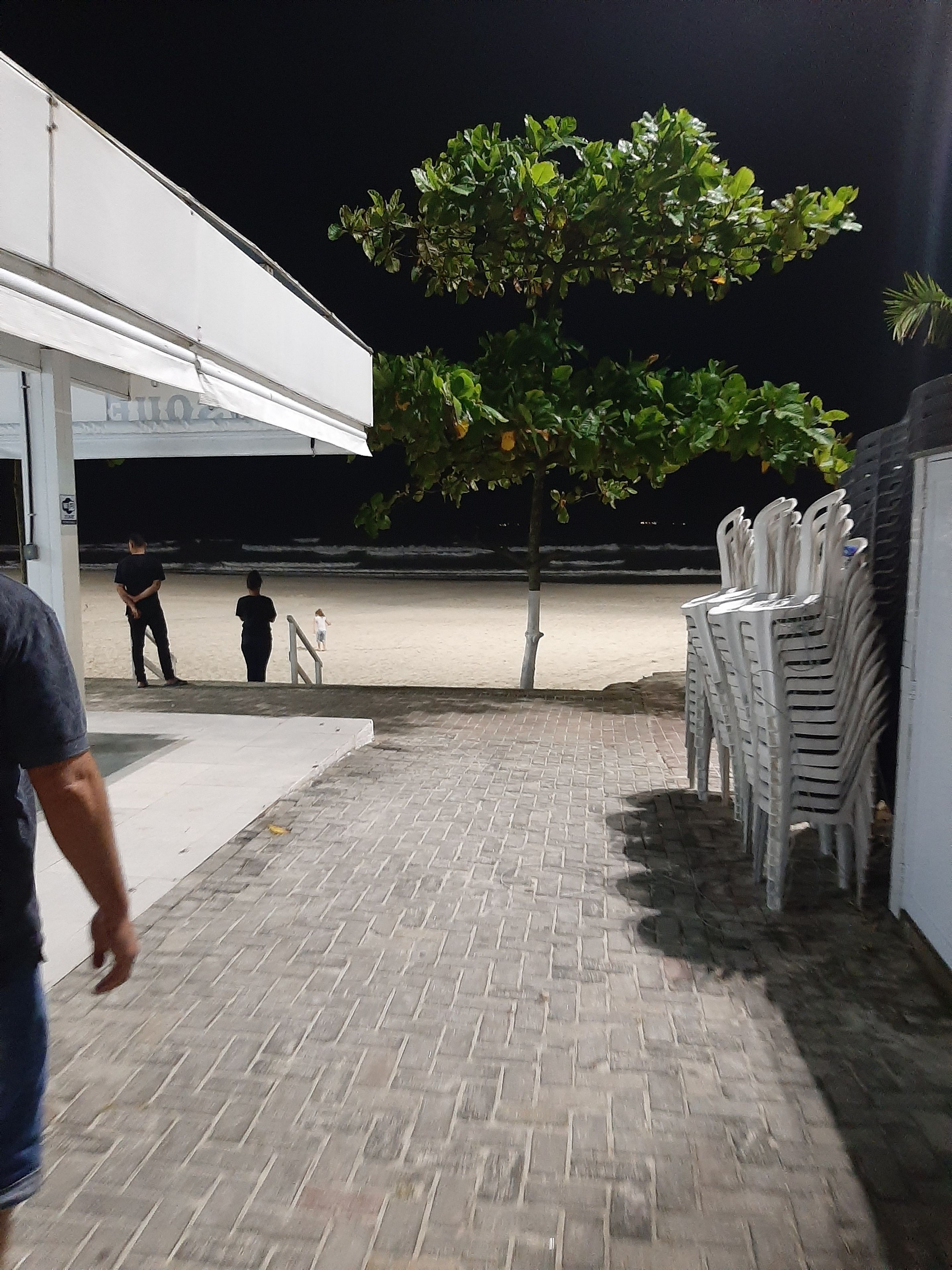 Uma área externa pavimentada com um edifício branco, uma árvore e uma pilha de cadeiras de plástico branco.