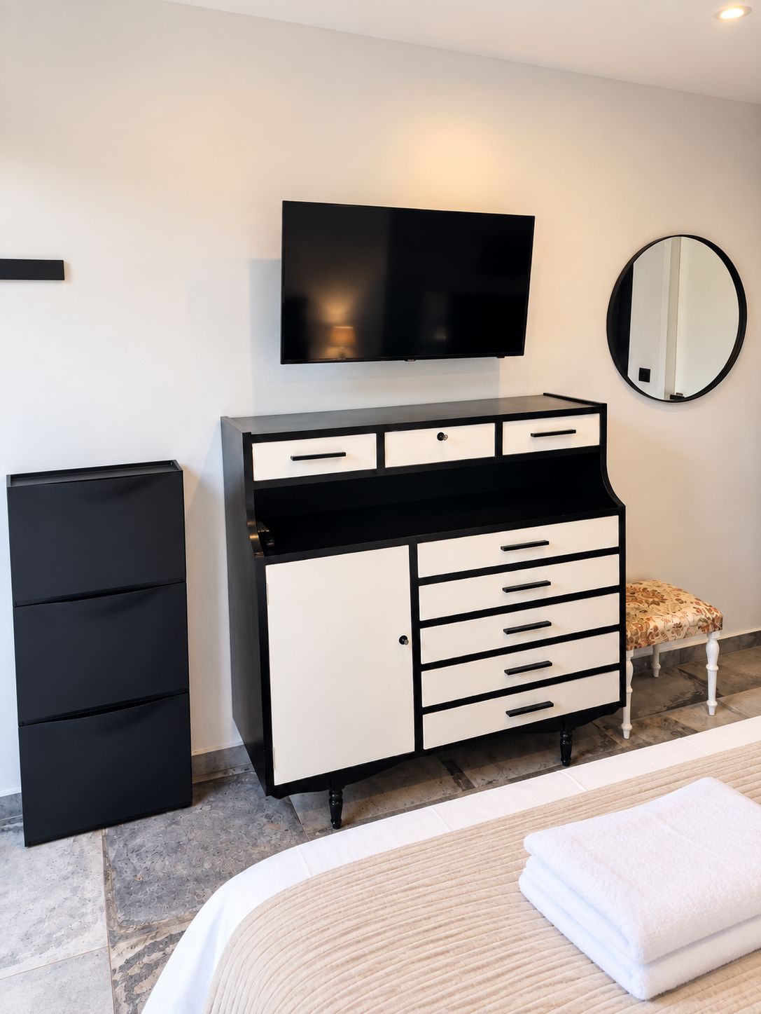 Une chambre d’hôtel avec un lit, une commode noire et blanche, un téléviseur, un miroir et un petit tabouret.