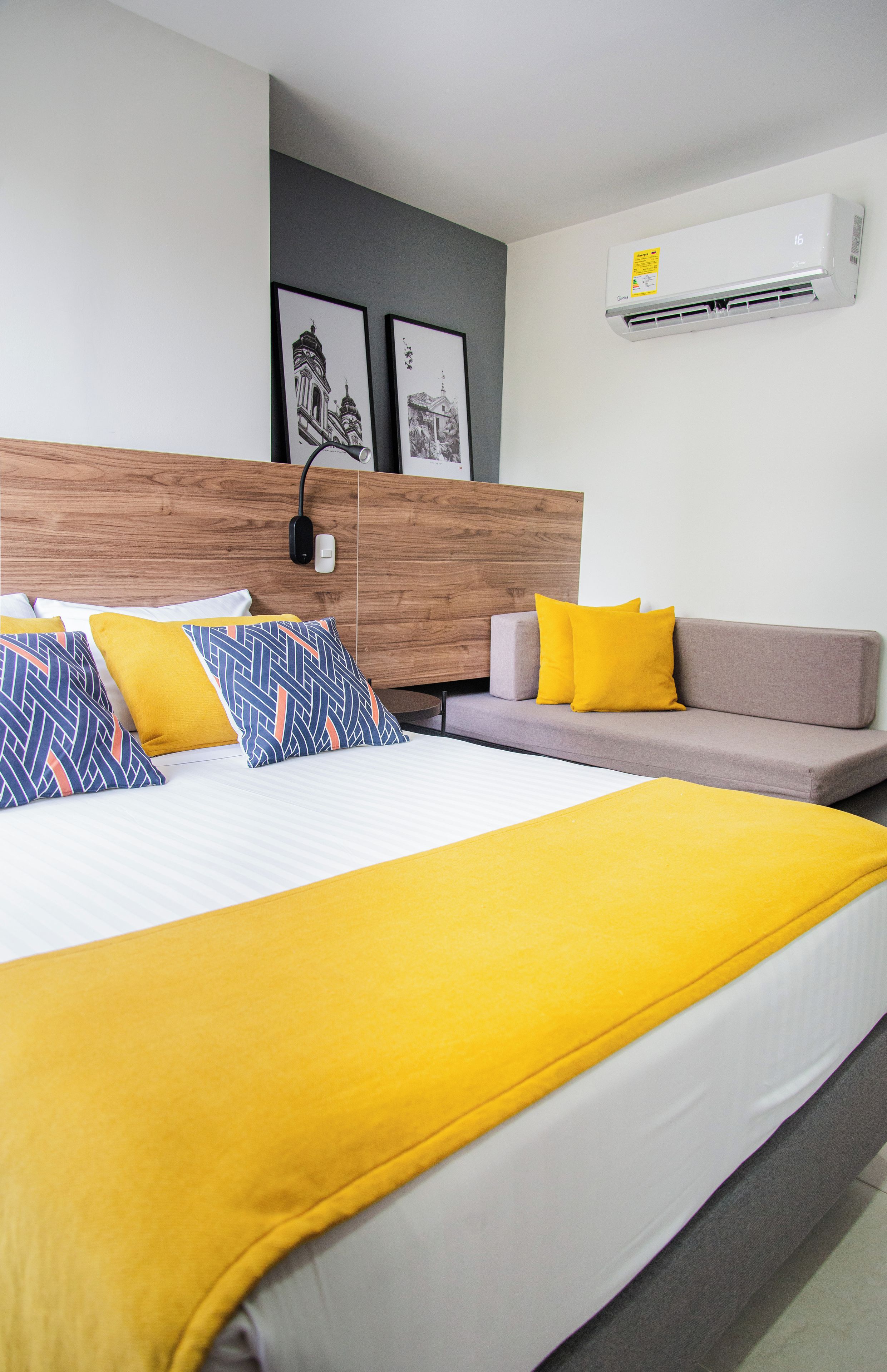 A hotel room with a bed, a sofa, a wall-mounted air conditioner, and two framed pictures.