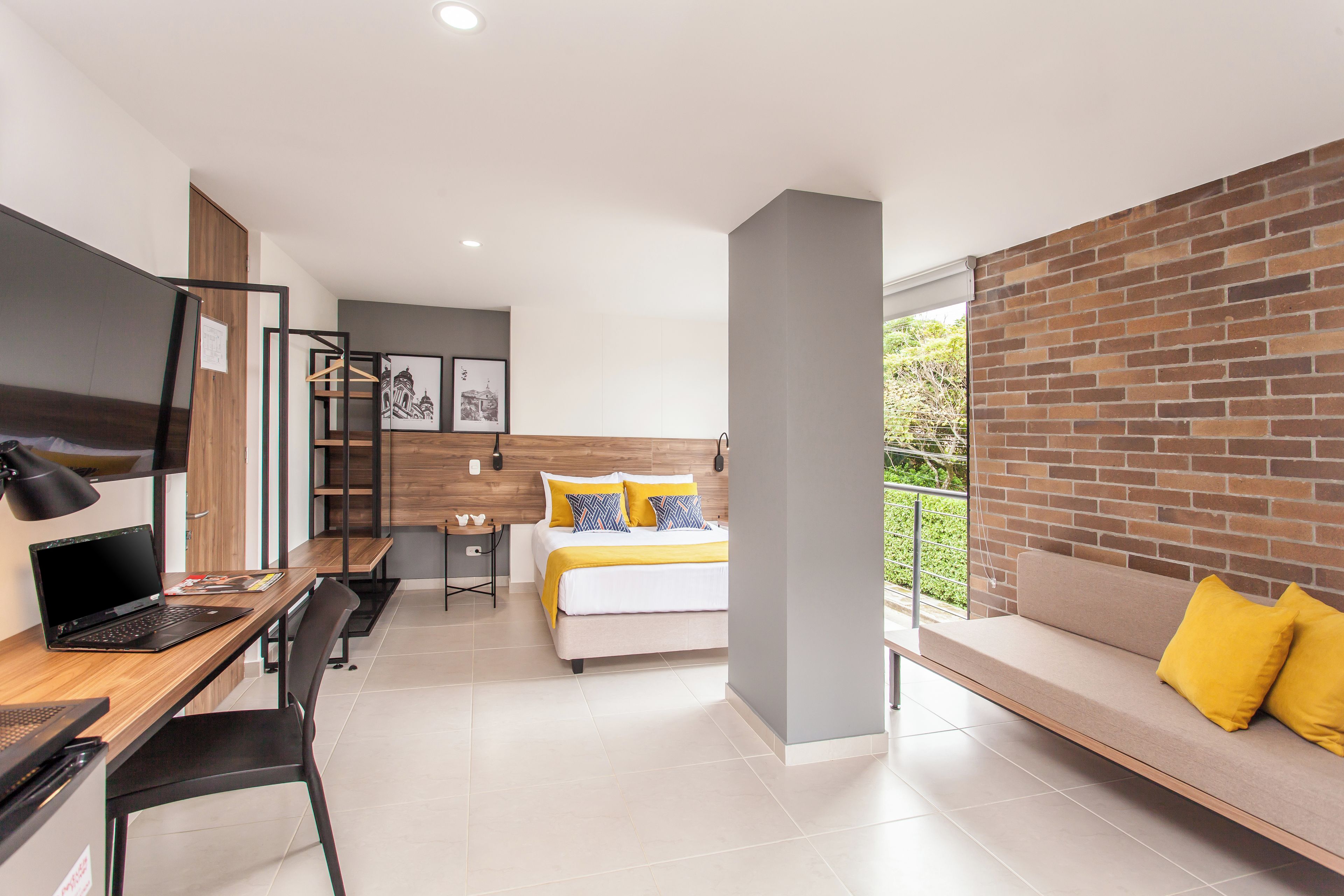 Habitación de hotel moderna con cama, escritorio con computadora portátil, una silla, un sofá y vista a zonas verdes por la ventana.
