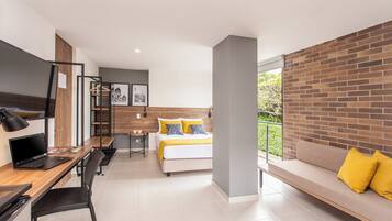 Habitación de hotel moderna con cama, escritorio con computadora portátil, una silla, un sofá y vista a zonas verdes por la ventana.