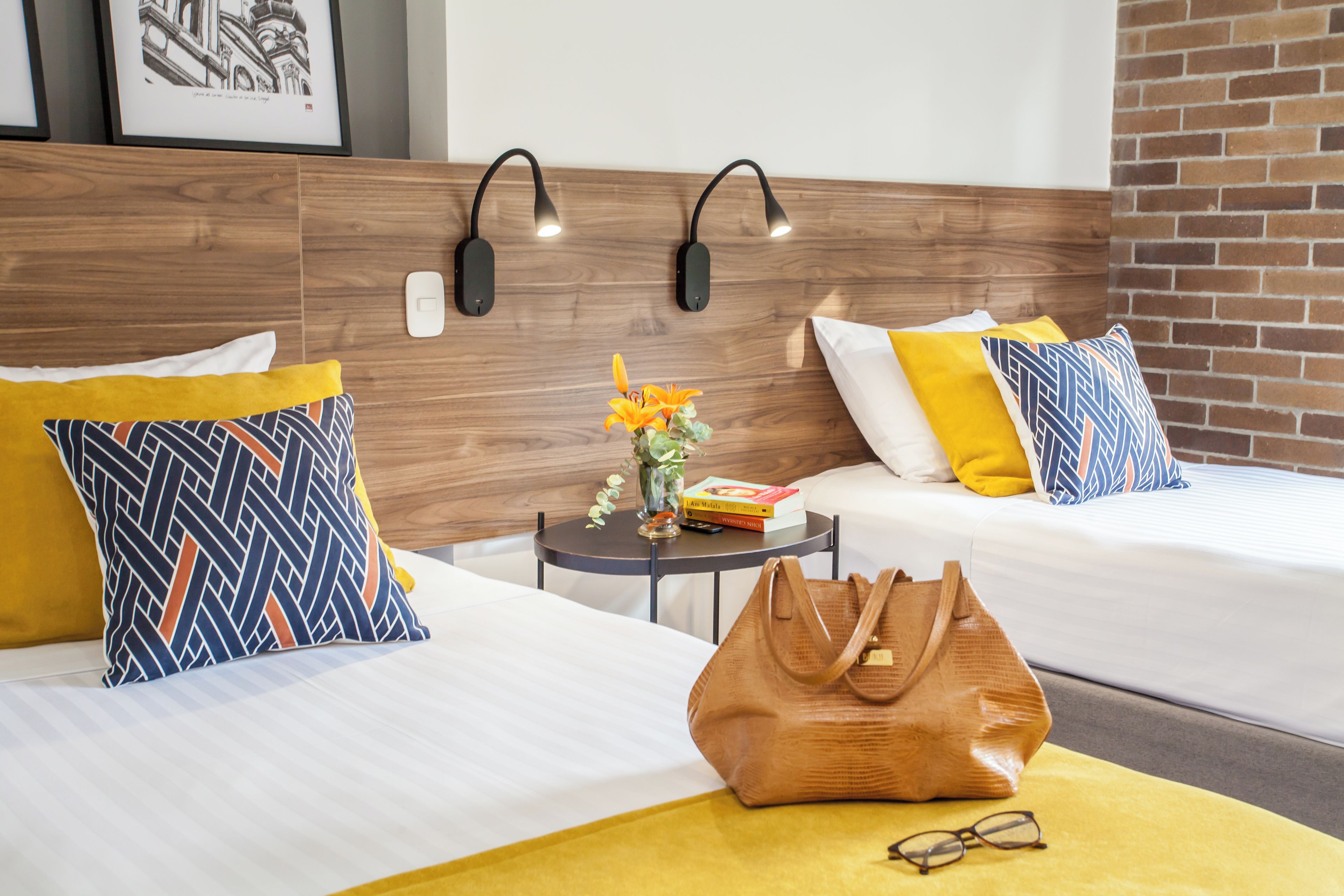 Hotellihuone, jossa on kaksi sänkyä, pieni pöytä, maljakko täynnä kukkia, kirja ja käsilaukku.