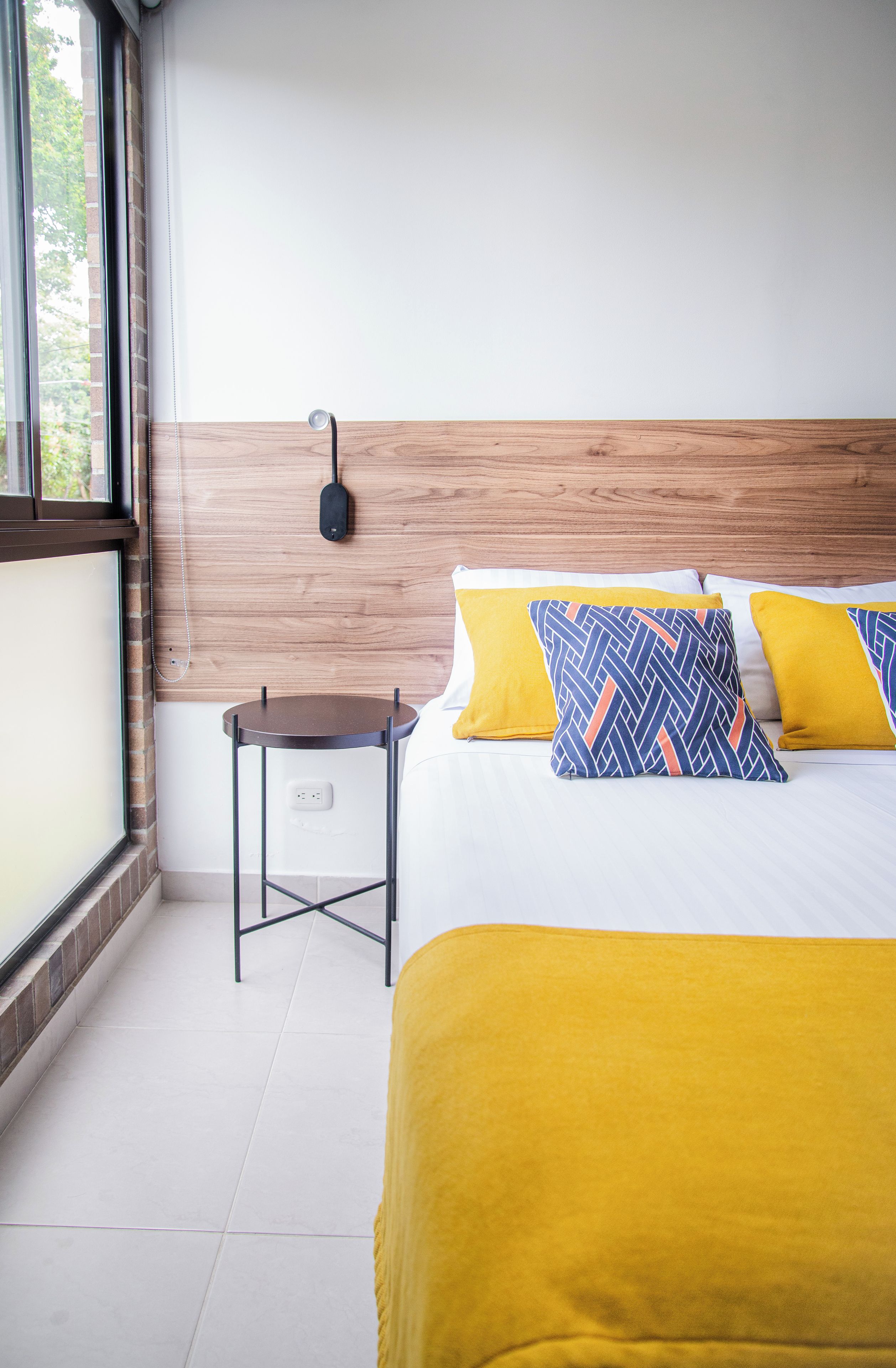 A hotel room with a bed, a bedside table, a lamp, and a window.