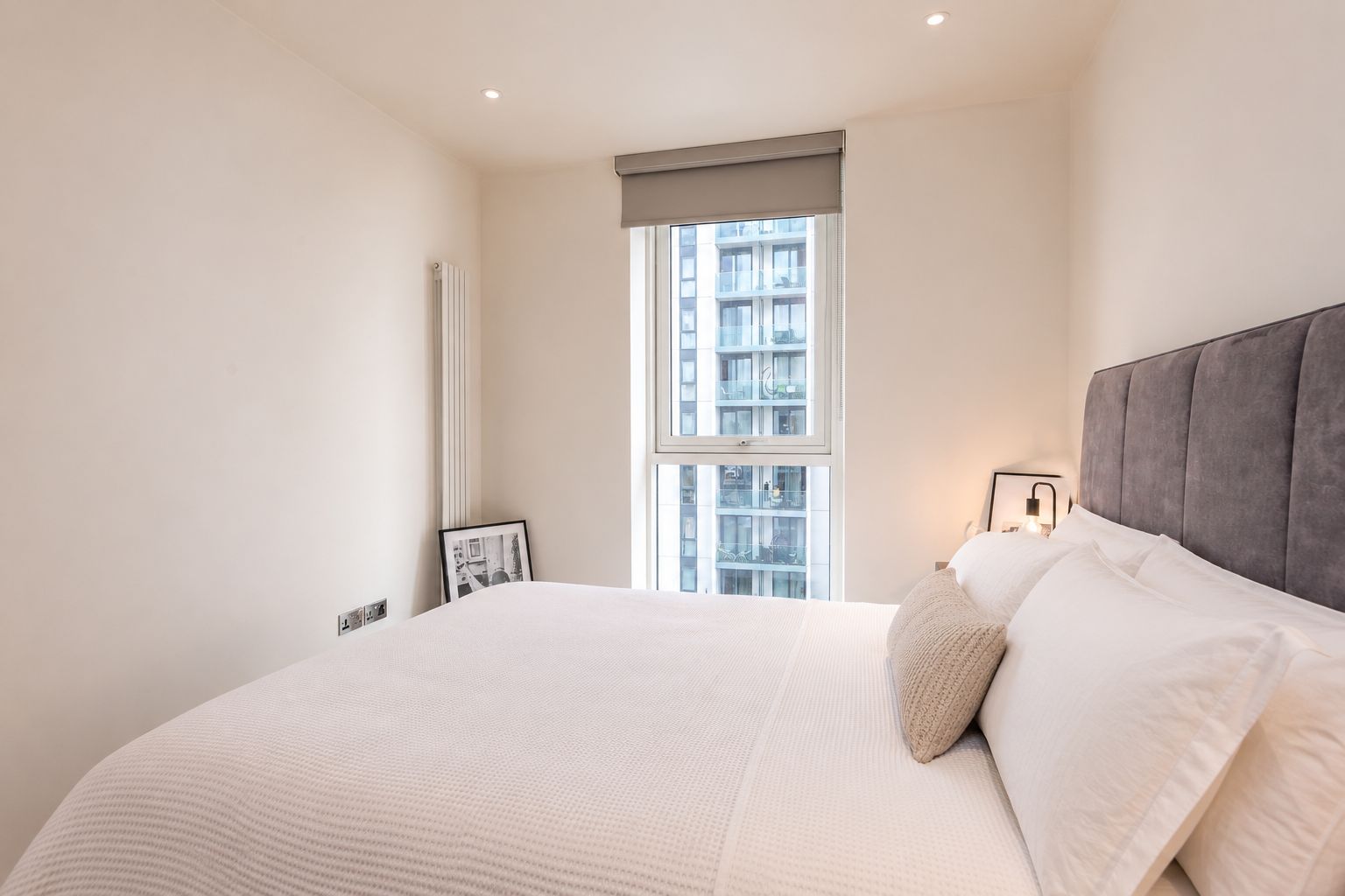 Un dormitorio con una cama grande, una ventana con vistas a edificios, un radiador e imagen enmarcada en la pared.