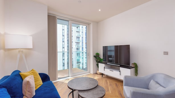A modern living room with a sofa, a TV, a small table, and a balcony with a view of high-rise buildings. - Green Rooms by Veeve (Salford)