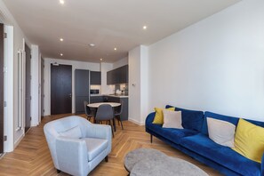 Una sala de estar moderna con un sofá, un sillón y un reposapiés. Se observa una cocina al fondo con una mesa de comedor y sillas.