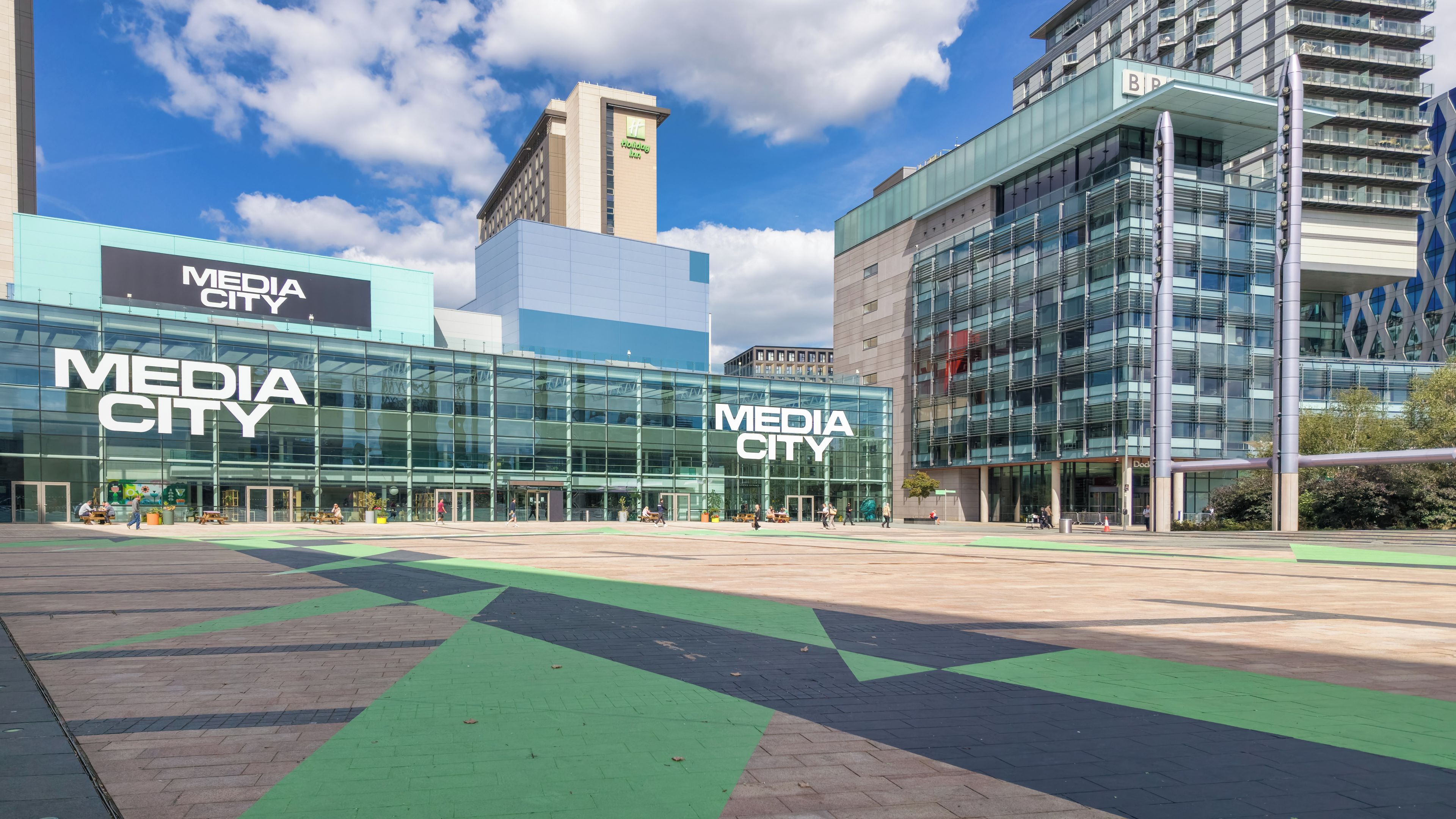 Um edifício moderno com a sinalização 'MEDIA CITY', com fachada de vidro e uma faixa de bicicleta verde.