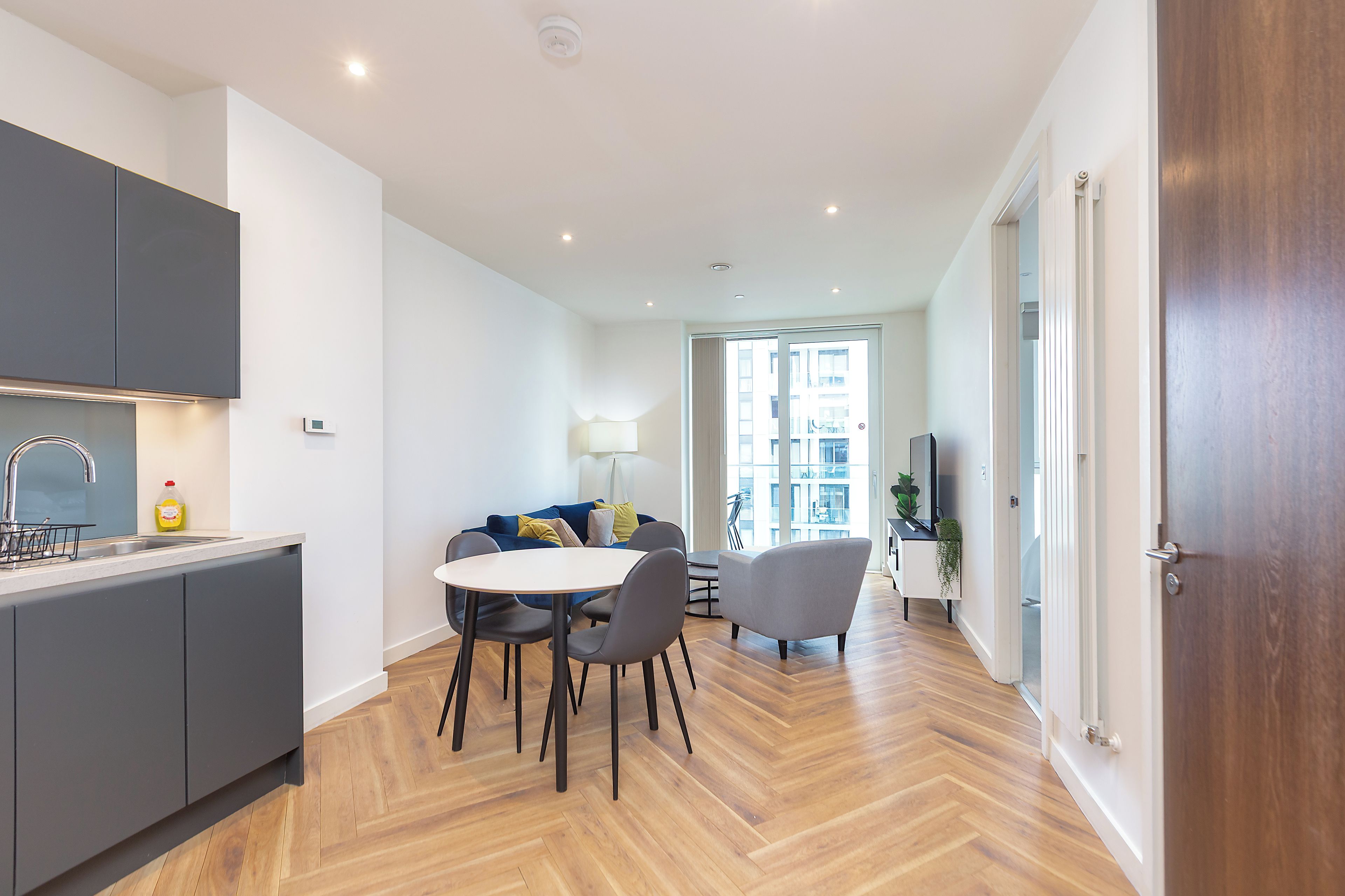 A modern kitchen with grey cabinets, a dining area with a round table and chairs, and a view into a living space with a sofa and a lamp.