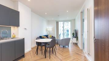 A modern kitchen with grey cabinets, a dining area with a round table and chairs, and a view into a living space with a sofa and a lamp.