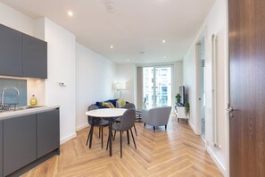 Une cuisine moderne avec des meubles de couleur grise, un coin repas avec une table ronde et des chaises, et une vue sur un espace de vie comprenant un canapé et une lampe.
