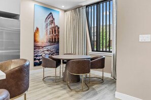 Ein Hotelzimmer mit einem runden Esstisch, vier Stühlen, einem großen Fenster mit Vorhängen und einem an der Wand befestigten Lichtschalter.