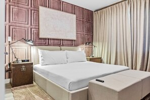 A neatly arranged hotel room with a large bed, bedside lamps, a bedside table, a suitcase, and a window with curtains. - 4BR house sleeps 10 near Magic Village Views (Kissimmee)