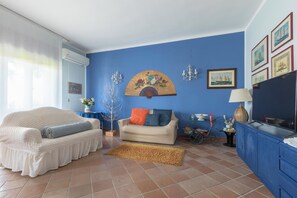 A living room with a blue wall, a beige sofa, a white armchair, a small table with a lamp, and a TV on a blue cabinet. - Villa Muse by BarbarHouse (Leporano)
