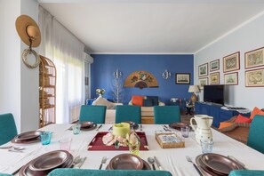 A dining area with a table set for four, blue upholstered chairs, a blue wall with framed artwork, and a blue sofa in the background. - Villa Muse by BarbarHouse (Leporano)