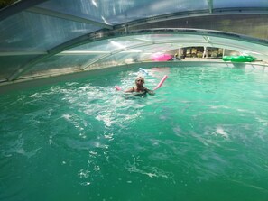 Zona de piscina con techo transparente, una persona nadando y juguetes inflables.