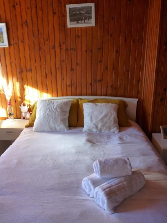 Een net opgemaakt bed met wit en geel beddengoed, een ingelijste foto aan de muur en een nachtkastje met een lamp en een vaas.