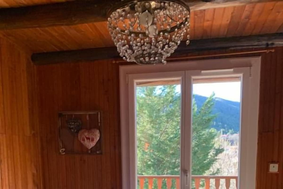 A room with a bed, a red chair, a chandelier, and a balcony with a view of trees and mountains.