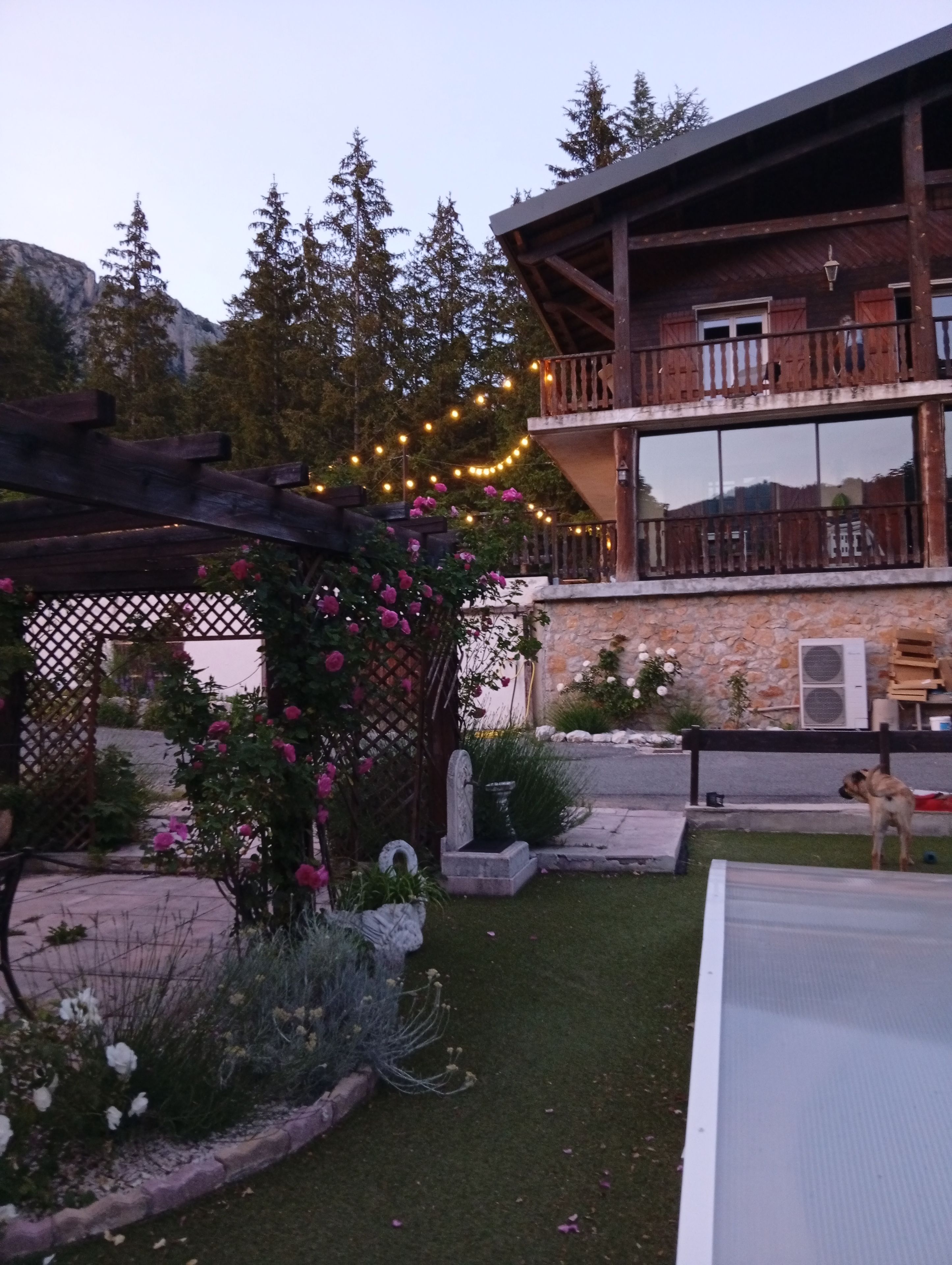 Un edificio de dos pisos con un balcón, una pared de piedra y un jardín con flores y una pérgola.