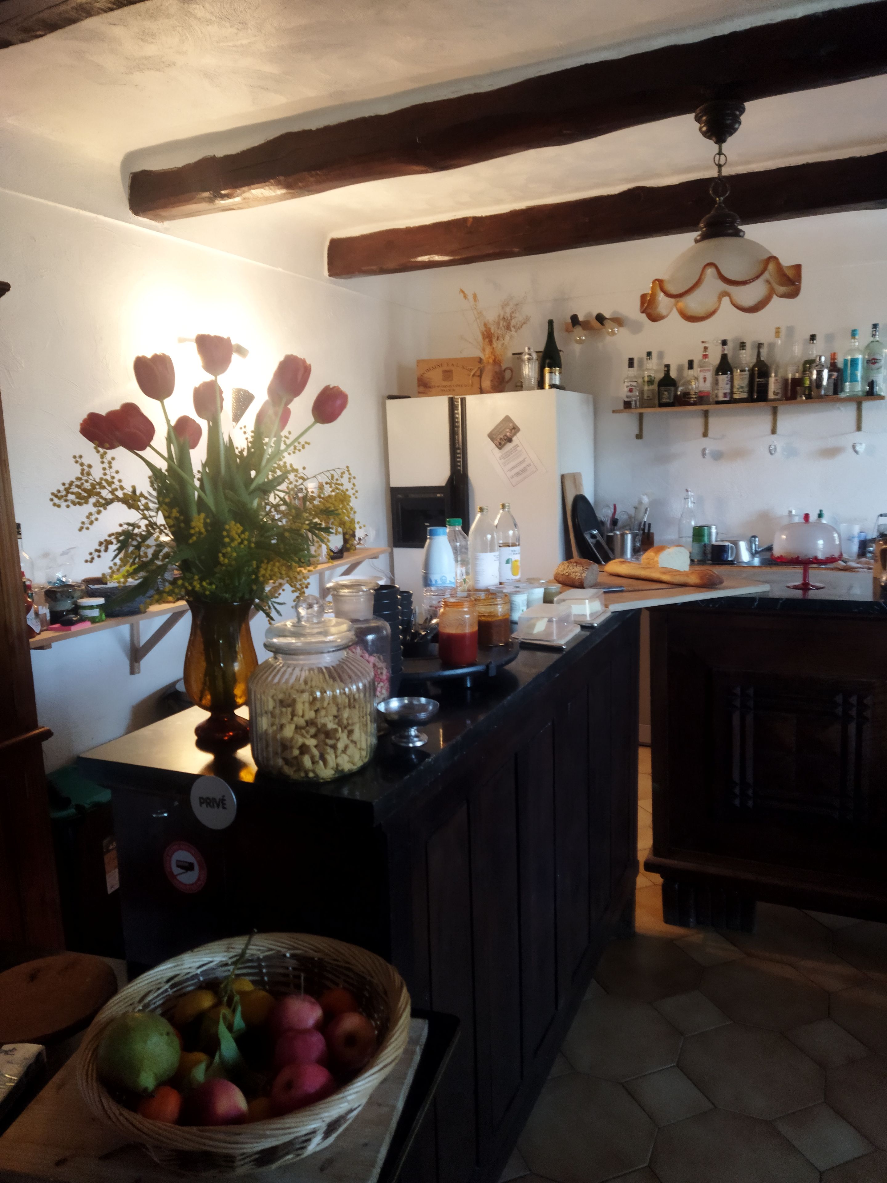 A kitchen with a wooden ceiling, a refrigerator, a countertop with various items, and a basket of fruit.