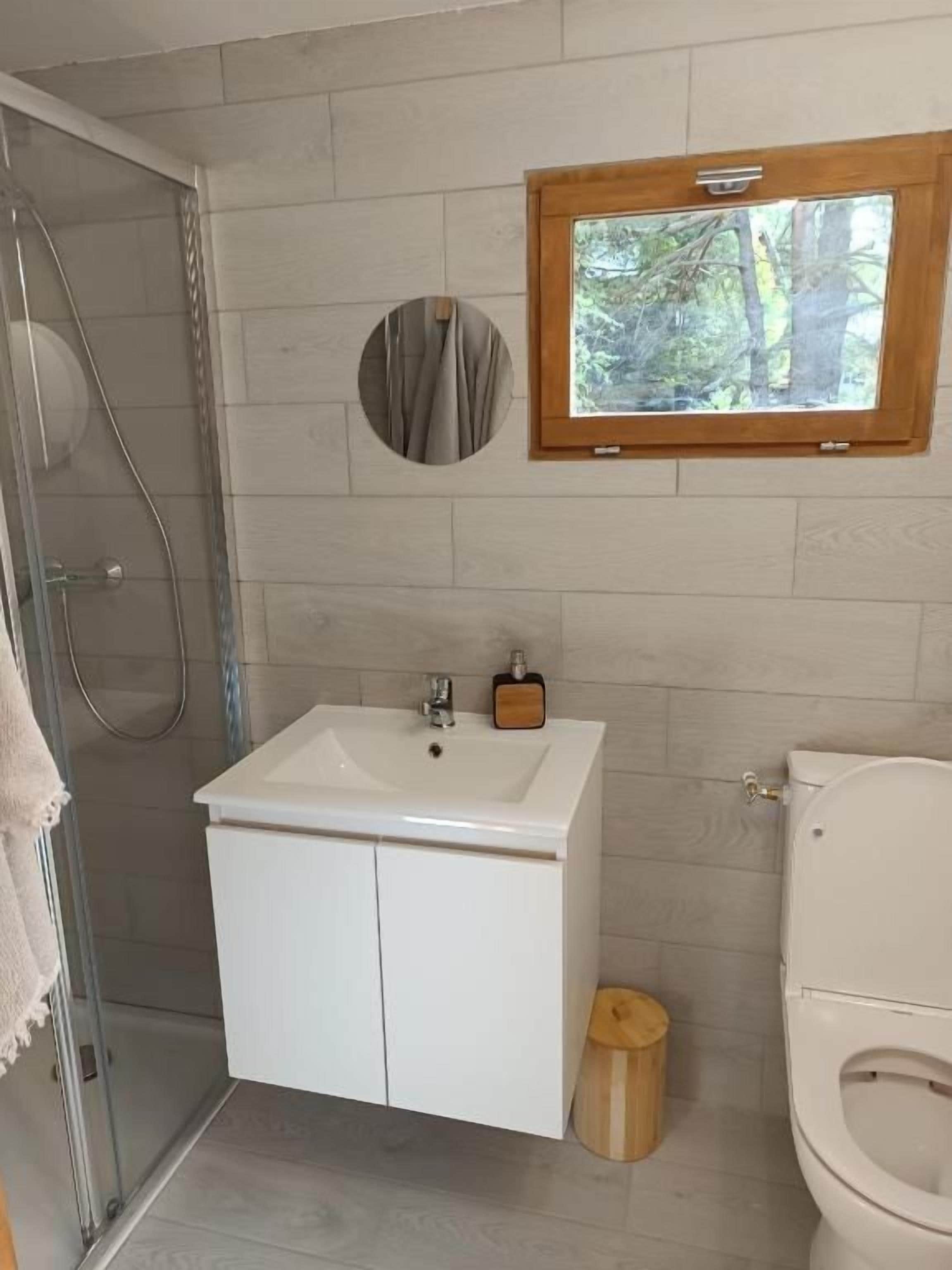 Un baño con ducha, lavamanos, inodoro y ventana con vistas a árboles.