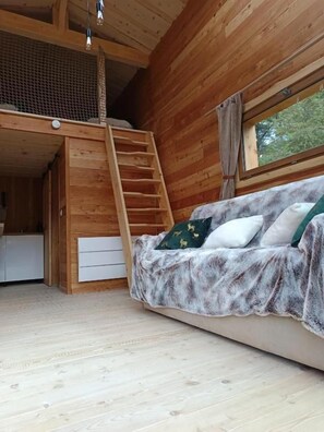 L’intérieur d’un chalet en bois, avec un lit en mezzanine, un canapé recouvert d’une couverture et une fenêtre donnant sur la verdure.