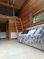Interior de una cabaña de madera con cama en altillo, sofá con manta y ventana con vistas a la vegetación.
