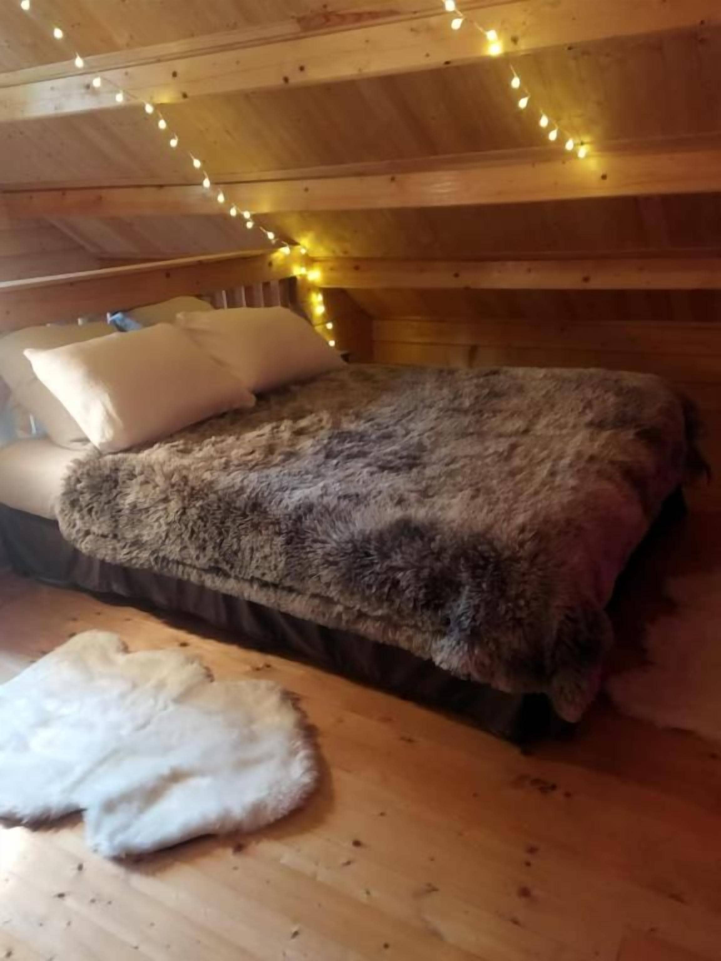 A wooden cabin interior with a bed, pillows, and a fur rug on the floor.