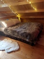 Interior de una cabaña de madera con una cama, almohadas y una alfombra de piel en el suelo.