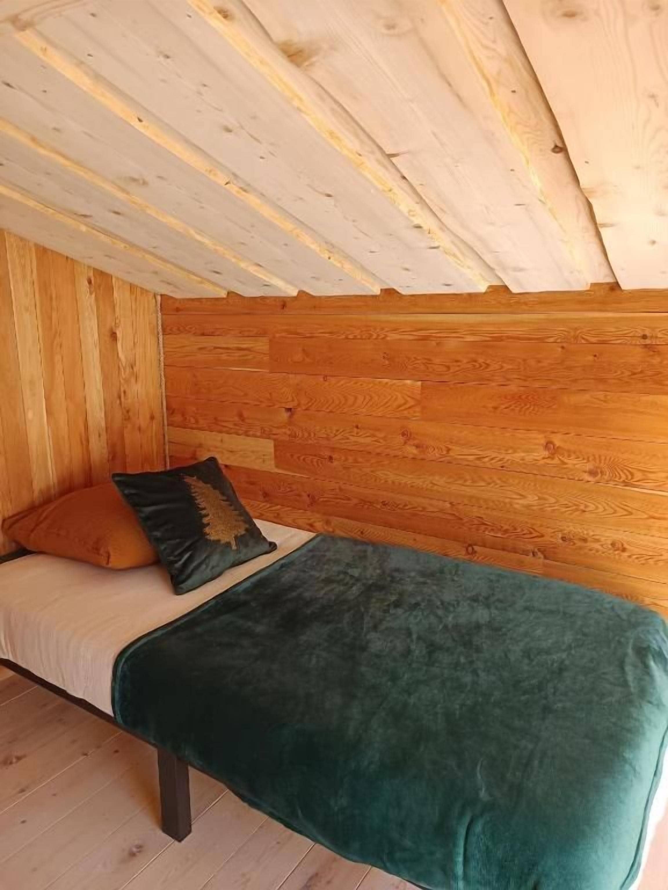Un lit simple avec une couverture verte et deux oreillers, dans une pièce aux murs en bois et au plafond en pente.