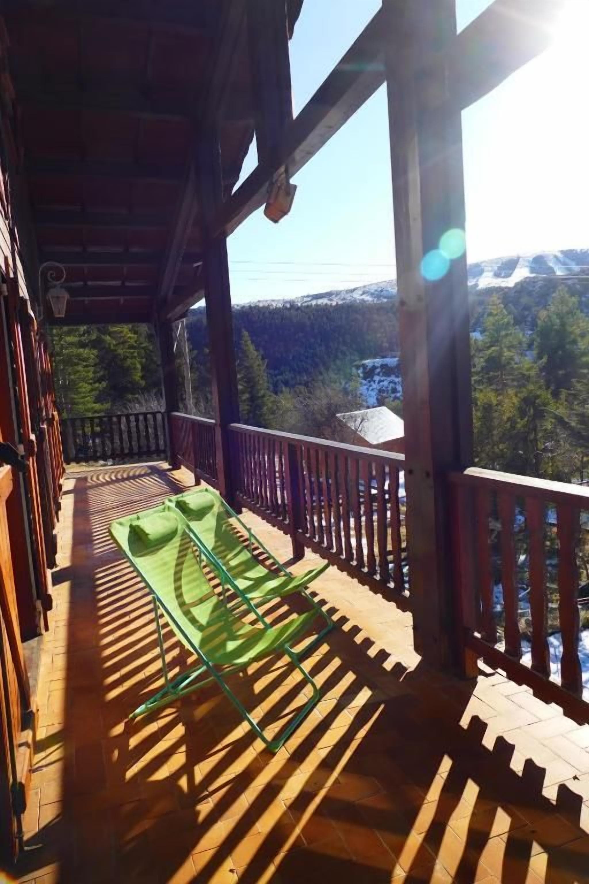Un balcón de madera con una silla verde, barandillas de madera y vistas a un área boscosa con montañas a lo lejos.