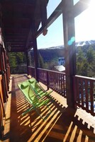 Un balcón de madera con una silla verde, barandillas de madera y vistas a un área boscosa con montañas a lo lejos.