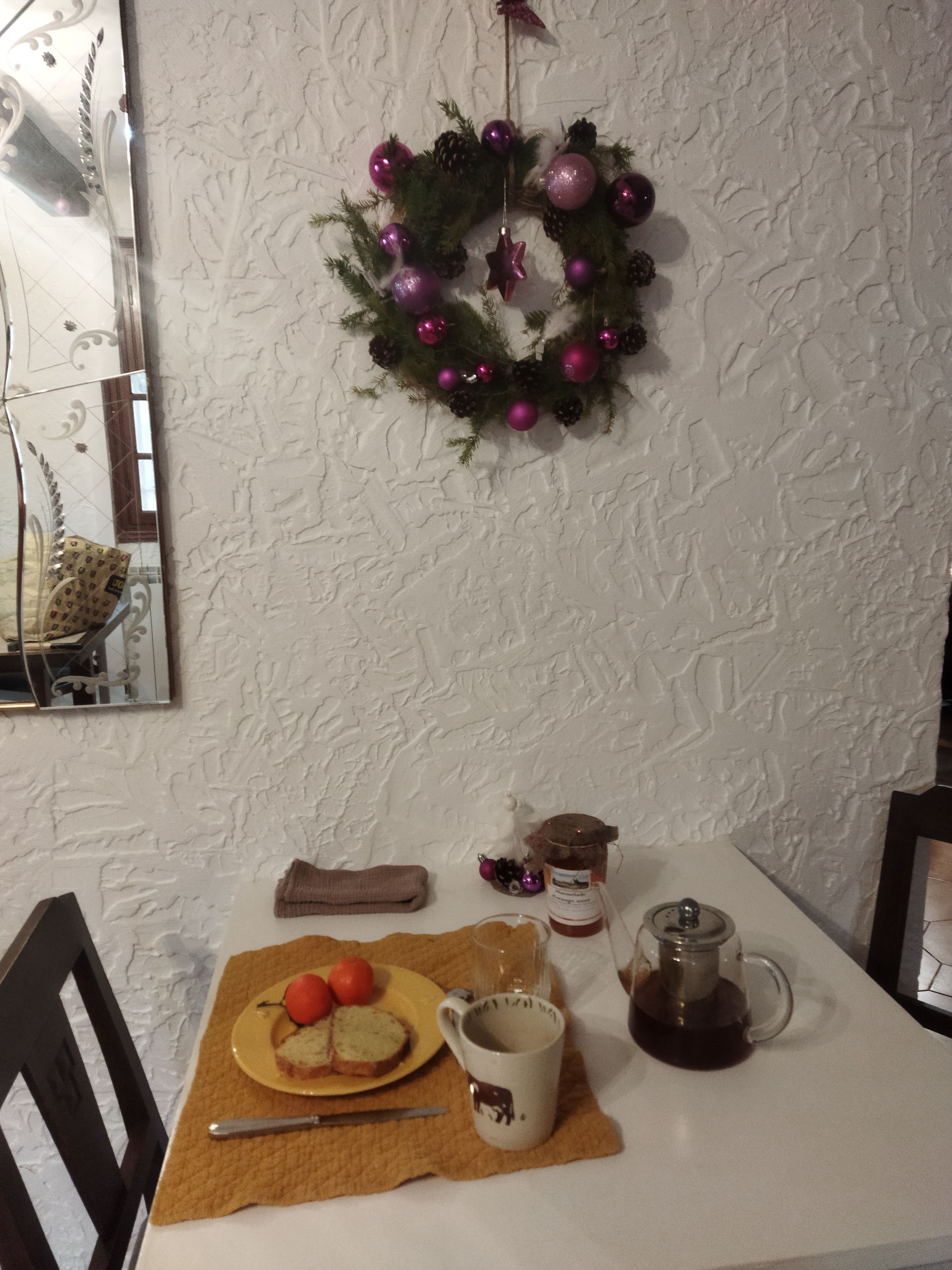 Ein Esstisch mit einer Speiseplatte, einer Tasse und einem Glas dunkler Flüssigkeit, möglicherweise Kaffee, an einer strukturierten weißen Wand mit einem dekorativen Kranz darüber.