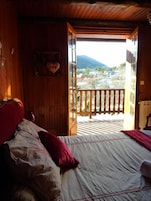 Un dormitorio con cama, cabecera de madera, ventana con vistas a una montaña y edificios, y un balcón con barandilla.