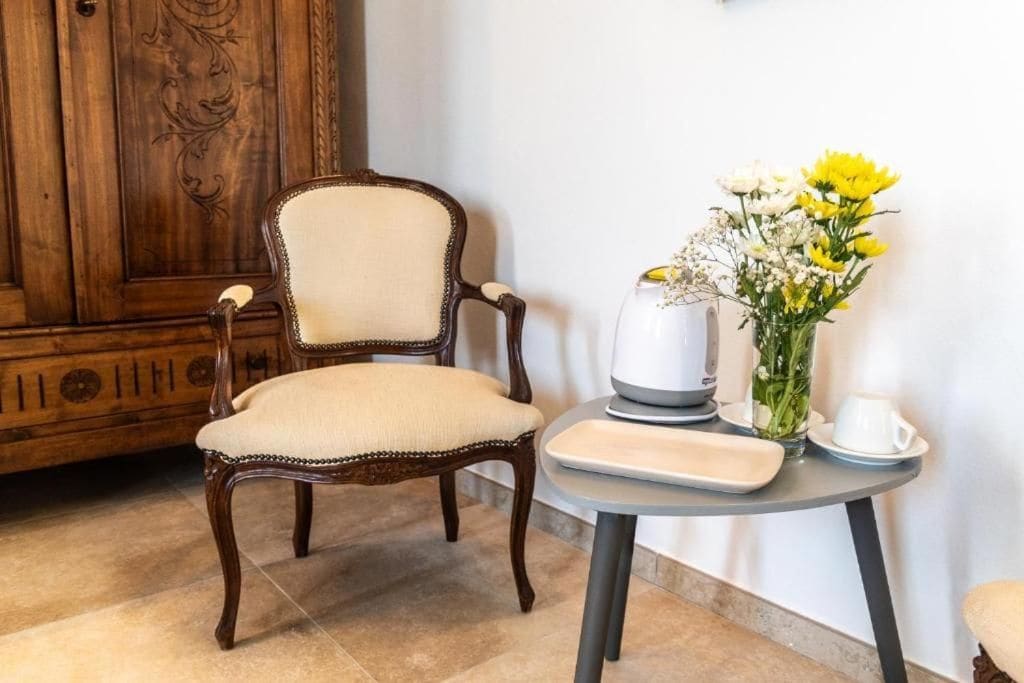 Une chaise en bois avec un siège et un dossier rembourrés, une petite table ronde sur laquelle se trouvent un pichet blanc et un vase de fleurs, et une tasse blanche posée sur la table.
