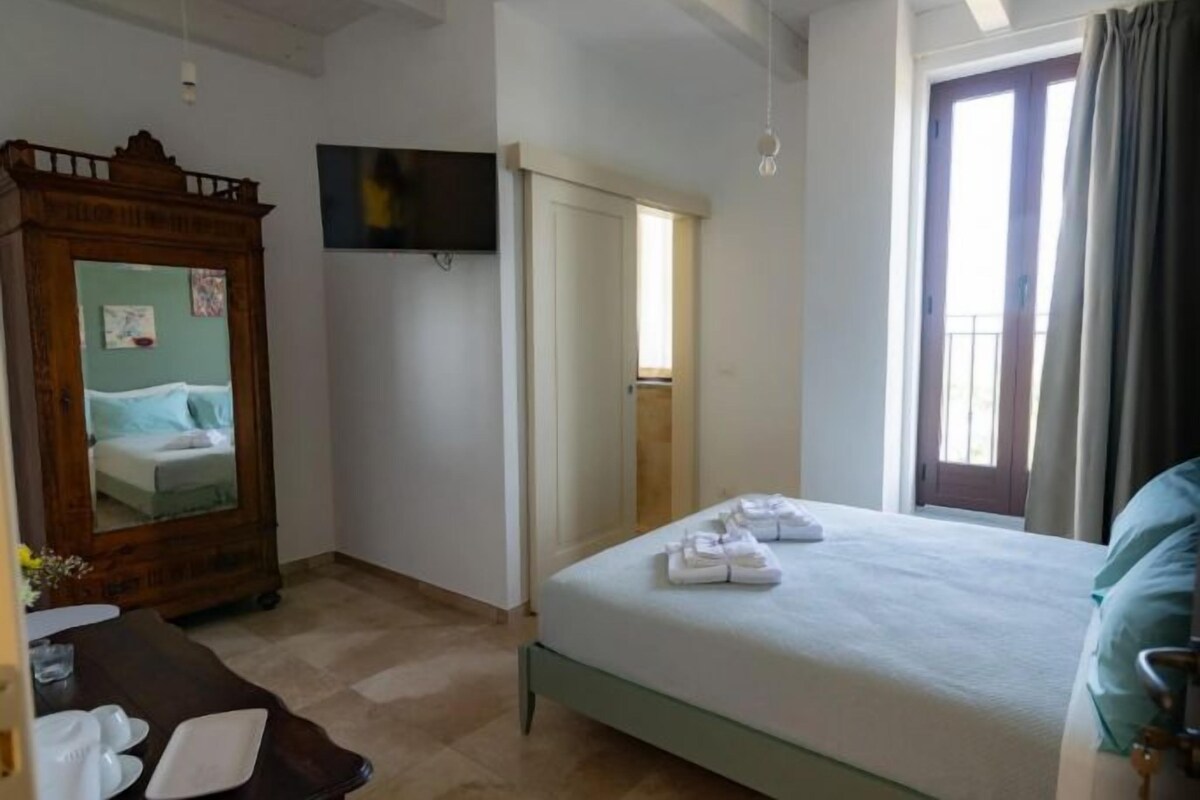 A hotel room with a bed, a wooden wardrobe, a flat-screen TV, and a balcony with curtains.