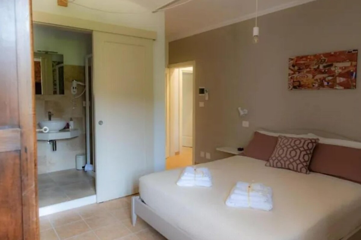 A bedroom with a bed, a bathroom visible through an open door, and a painting on the wall.