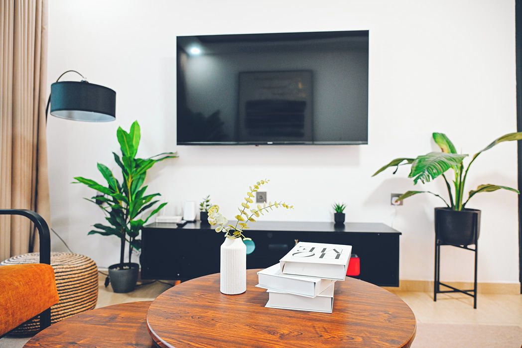 A modern living room with a round wooden table, a white vase with flowers, a stack of books, a black TV on the wall, and potted plants.