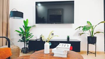 A modern living room with a round wooden table, a white vase with flowers, a stack of books, a black TV on the wall, and potted plants.