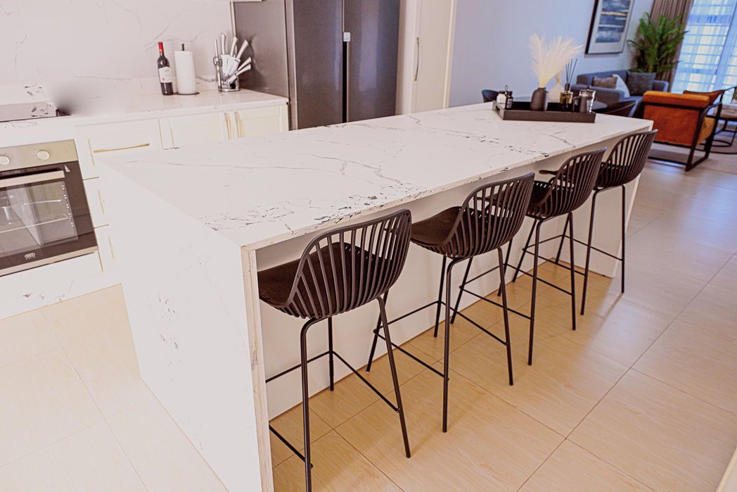 A modern kitchen with a marble countertop, four black chairs, a refrigerator, and a dining area with a plant.
