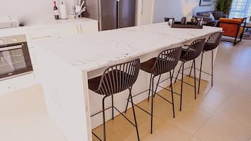 A modern kitchen with a marble countertop, four black chairs, a refrigerator, and a dining area with a plant.
