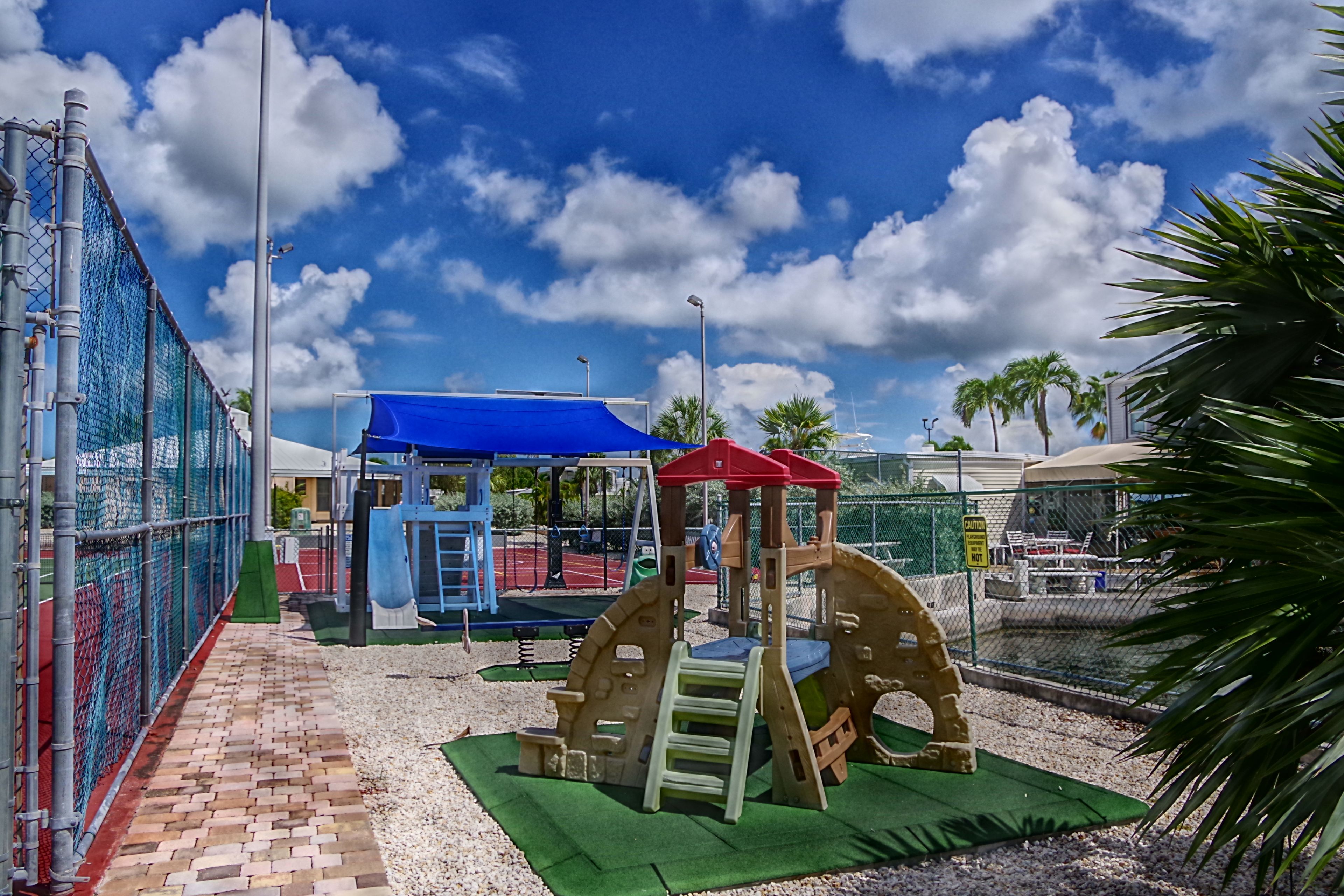A playground with various play structures, including a climbing frame and a swing set, surrounded by a fence and palm trees.