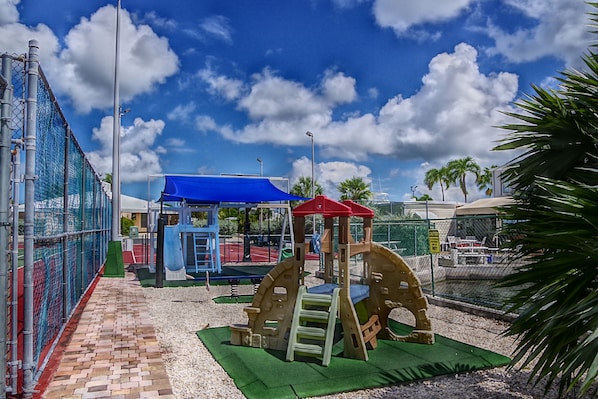 A playground with various play structures, including a climbing frame and a swing set, surrounded by a fence and palm trees.