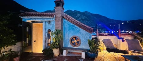 A building with a tiled roof, a door, and a circular window. There are potted plants and a bench outside.