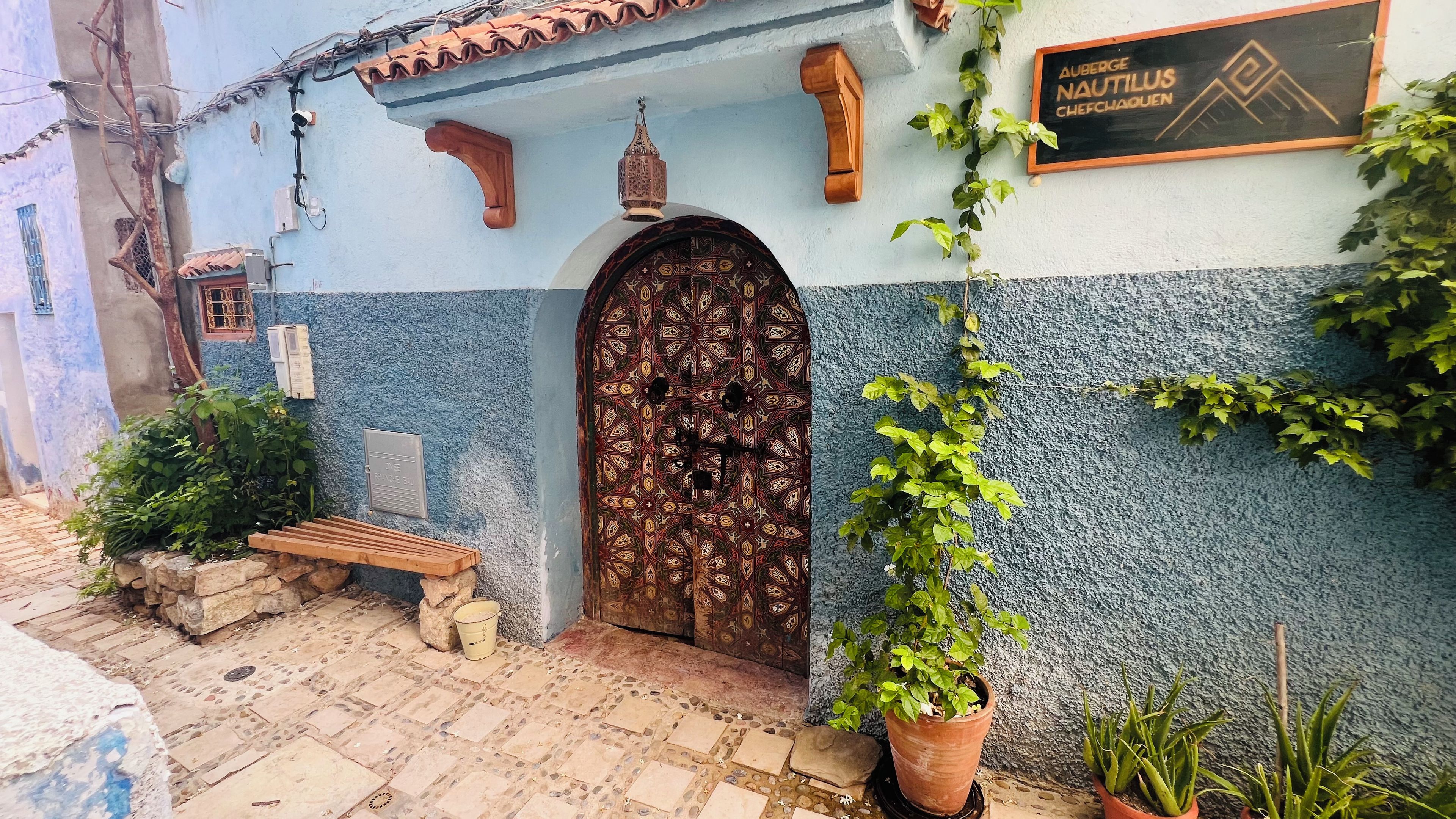 Um edifício tradicional com porta estampada, um banco de madeira e plantas em vasos.