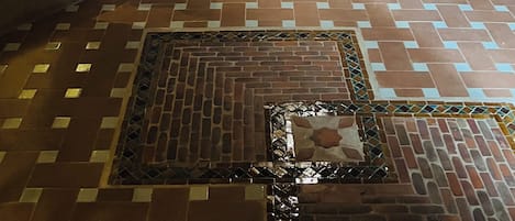 A room with a brick wall, a window with a decorative arch, a cat resting on a bench, and a patterned floor.