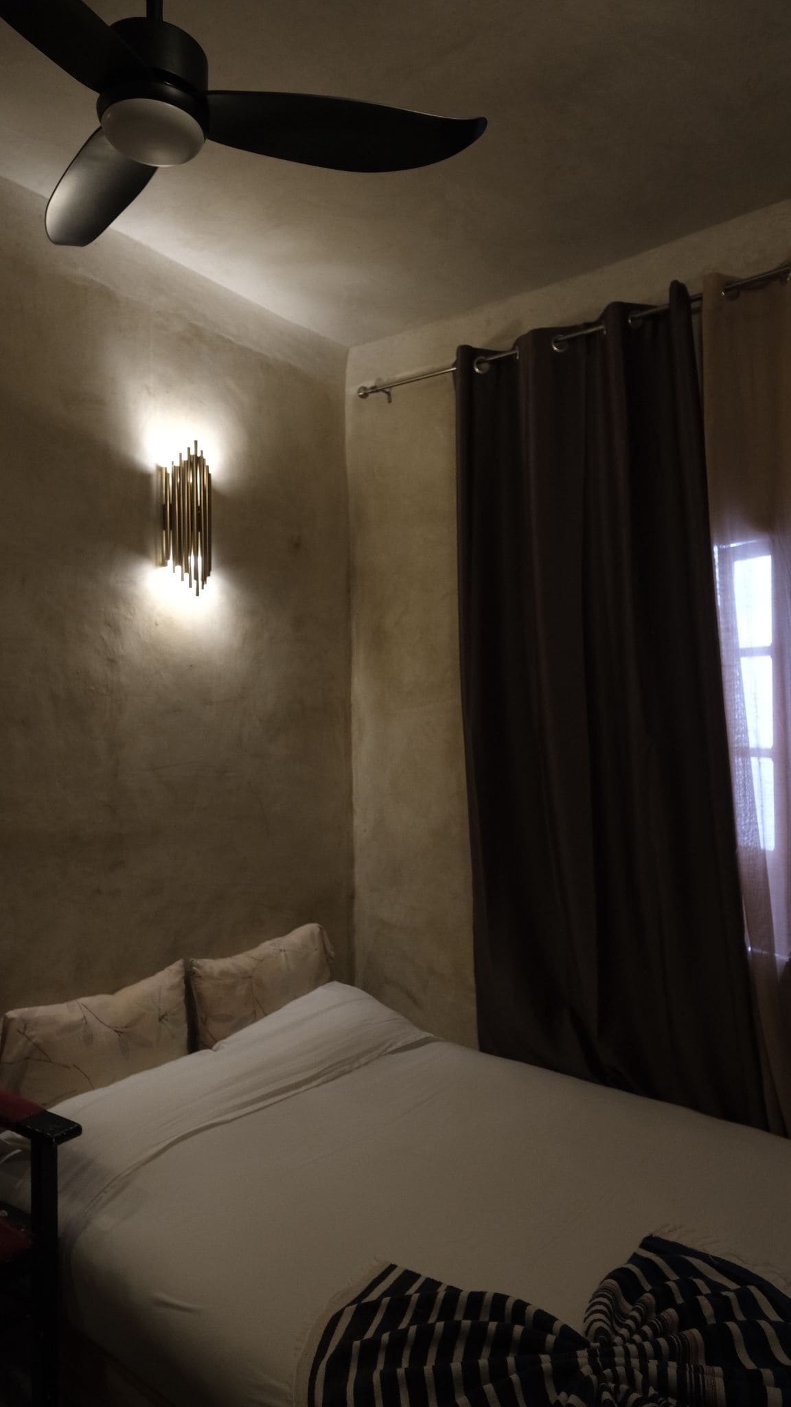 Un dormitorio con cama, ventilador de techo, luz empotrada en la pared y ventana con cortinas.