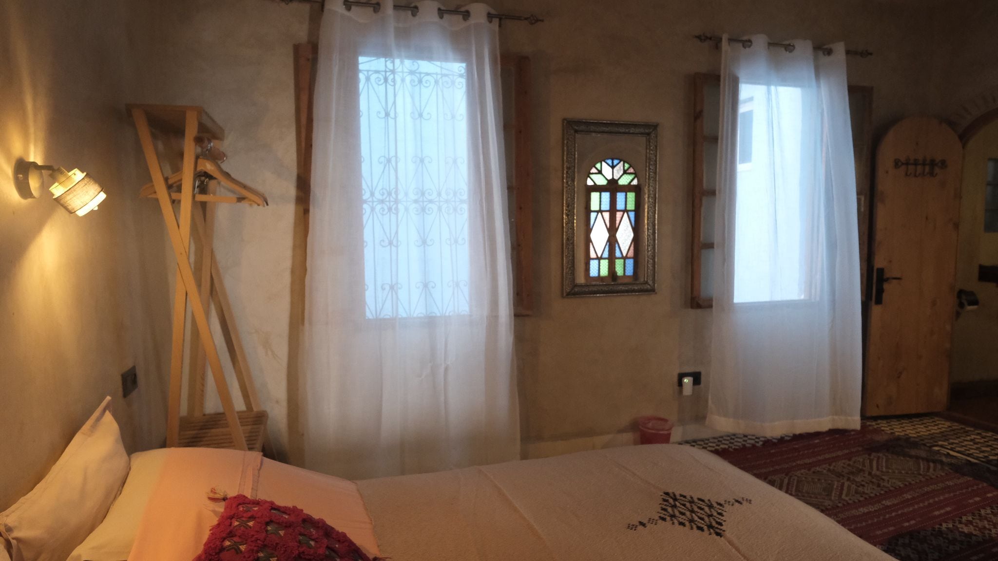 A room with a bed, a wooden chair, a window with a stained glass design, and a door with a wooden panel.